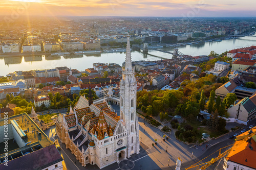 Budapest, Hungary - Aerial drone view of the beautiful Matthias Church at Castle district with a warm summer sunrise. Fisherman's Bastion and Szechenyi Chain bridge at background © zgphotography