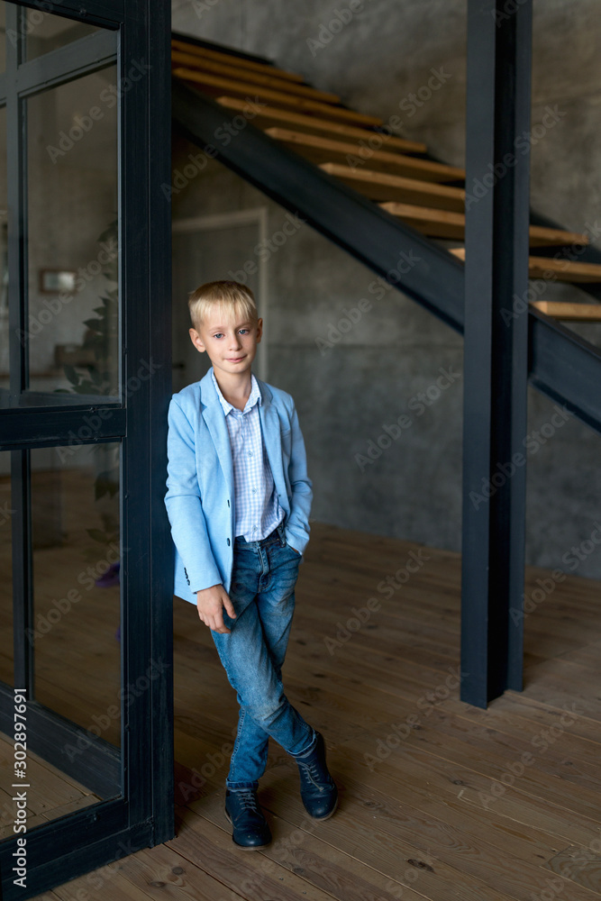 The blonde teenager, in a stylish shirt and jeans, stands in a loft-style apartment and smiles broadly at the camera