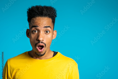 Amazed mixed race man shocked, saying WOW. Handsome african american guy with afro hair surprised to camera over blue background. Copy space.