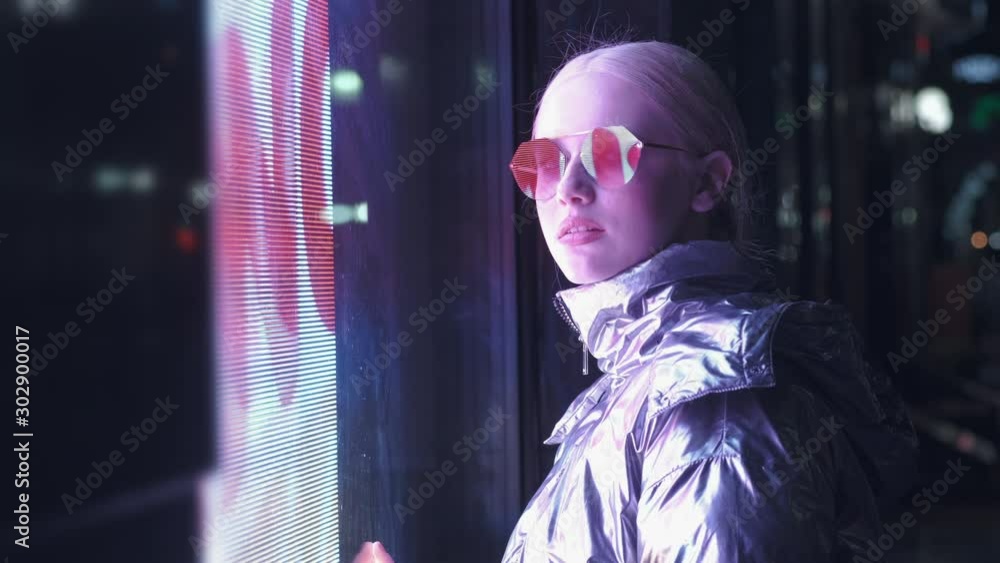 A young girl teenager model stands in neon lights. Millennial sad pretty girl with unusual hairstyle near glowing neon of the city at night. Nightlife atmosphere of girl looking to camera A young girl teenager model stands in neon lights. Millennial sad pretty girl with unusual hairstyle near glowing neon of the city at night. Nightlife atmosphere of girl looking to camera