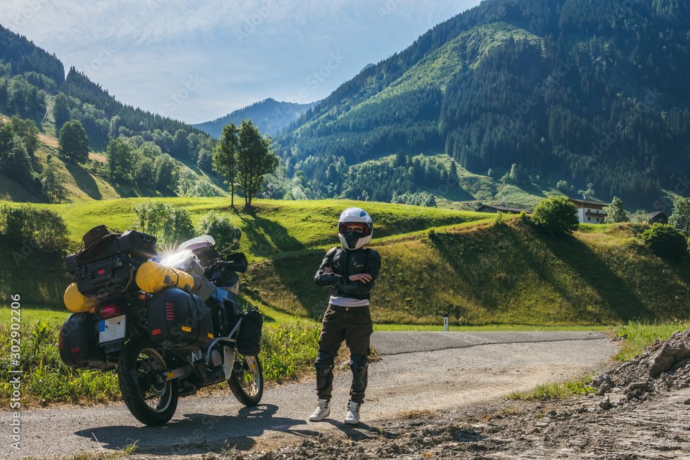 Fototapeta premium Girl in full body protection, turtle armor, knee pads and helmet, motorcycle for traveling with large luggage, on a background of green mountains, copy space, rady to ride