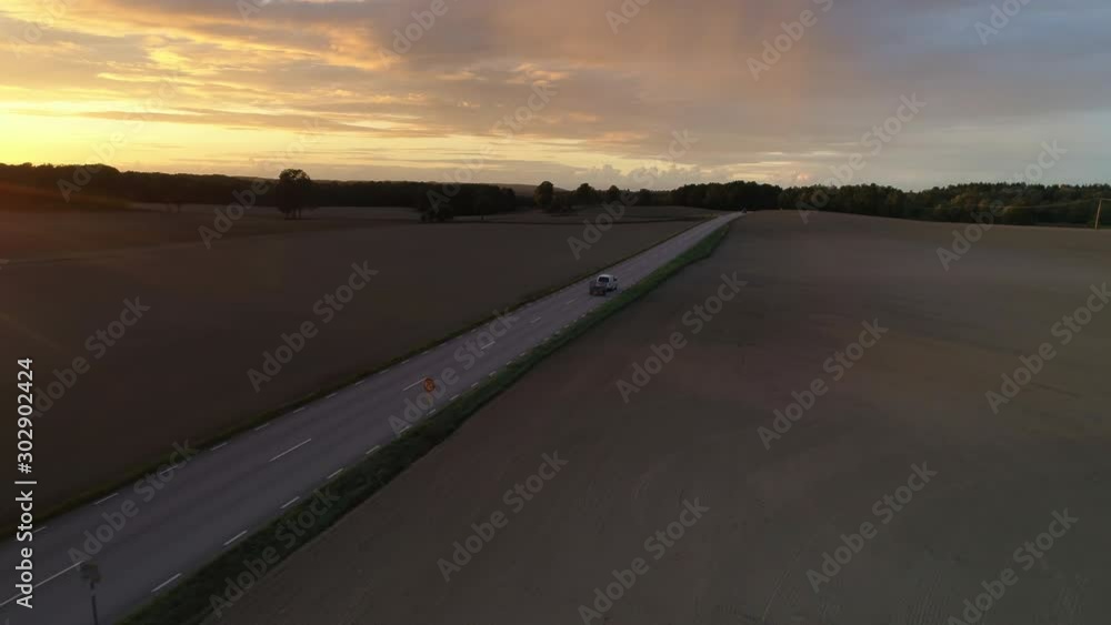 Country road and fields at sunset aerial view. Drone shot flying backwards over cars driving on road Country road and fields at sunset aerial view. Drone shot flying backwards over cars driving on road