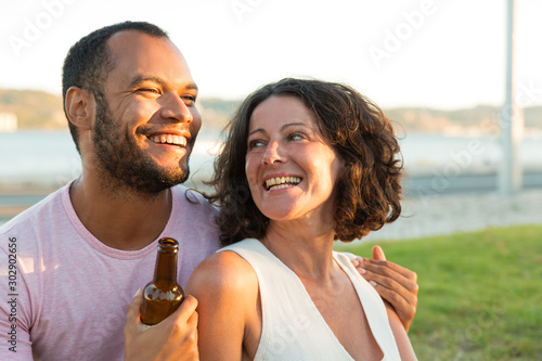 Happy relaxed couple drinking beer, and chatting outdoors. Man and woman sitting on grass, hugging, holding beer bottle and laughing. Dating outdoors concept