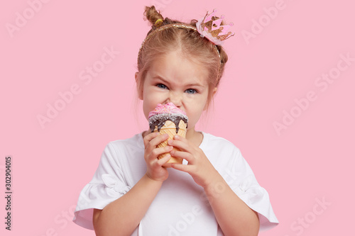 Cute child girl with pleasure eats tasty ice-cream on a pink studio background. Licks with closed eyes. The concept of baby food and a happy childhood