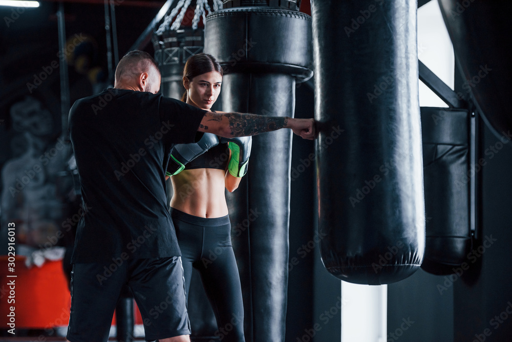 Young tattooed boxing coach teaches young woman in the gym