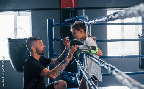 Conception of assistance. Young tattooed boxing coach teaches cute little girl in the gym