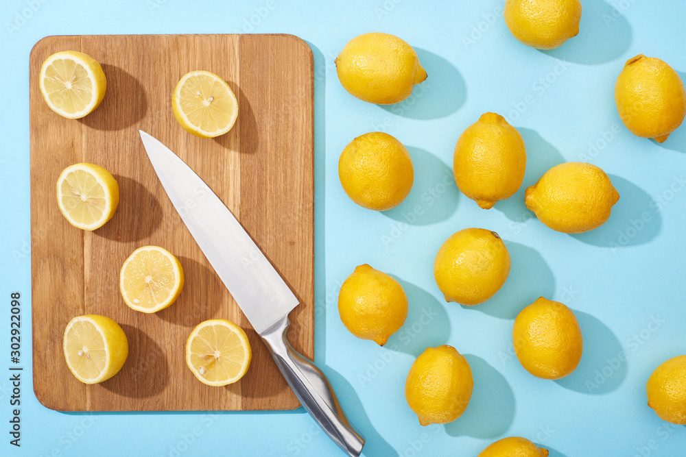 top view of ripe yellow cut lemons on wooden cutting board with knife on blue background near whole lemons top view of ripe yellow cut lemons on wooden cutting board with knife on blue background near whole lemons