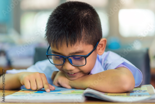 Asian elementary school boy in a white school uniform and wearing glasses, is reading a book in the classroom.