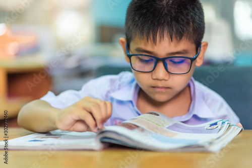 Asian elementary school boy in a white school uniform and wearing glasses, is reading a book in the classroom.