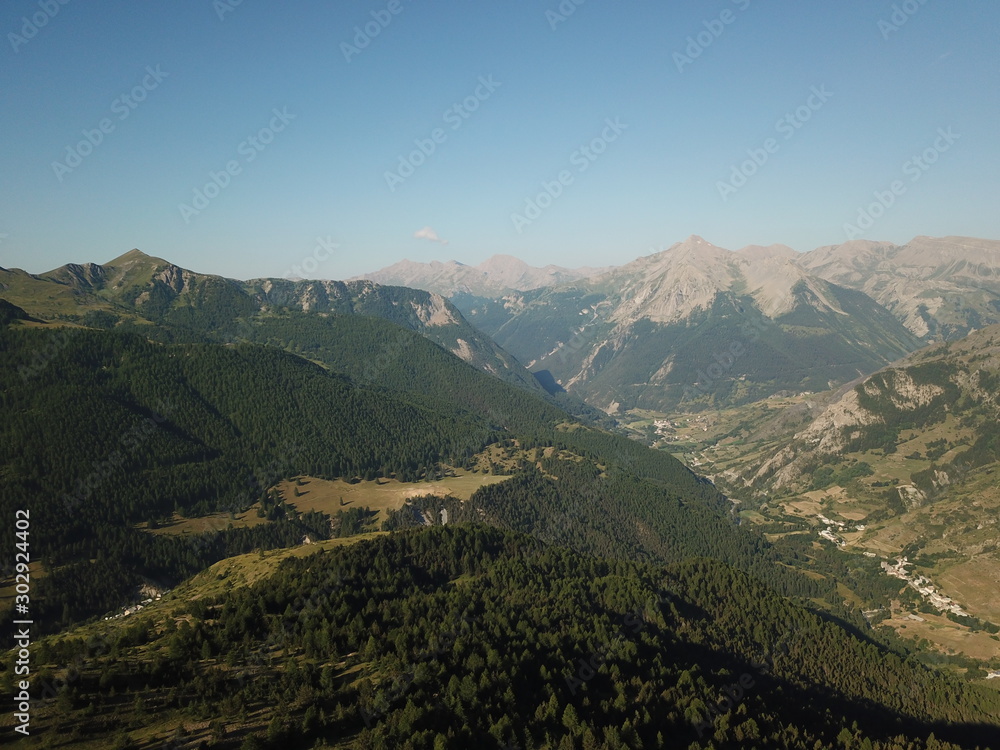 Obraz premium vue aérienne de vallée de haute montagne sous grand soleil avec sapins verts et village au loin et ciel bleu