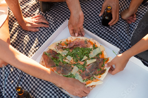 Group of people taking pizza slices from box outdoors. Top view of pizza in box on plaid, beer bottles and human hands. Outdoor dinner concept