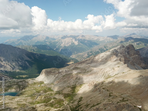 vue aérienne en altitude de sommets et d'une vallée de montagne verte et rocheuse avec ciel bleu et nuages cotonneux et percées de soleil sur les flancs de montagnes et avec un lac d'altitude
