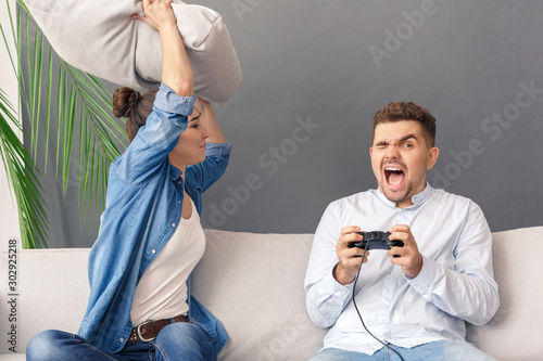 Relationnship Concept. Young couple woman hitting with pillow man playing game console screaming sitting on sofa studio isolated on grey photo