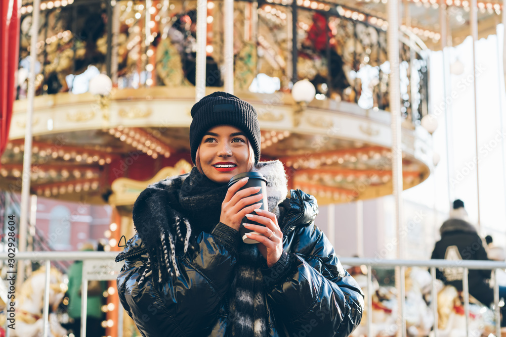 Beautiful woman standing by illuminated Christmas carousel with takeaway coffee
