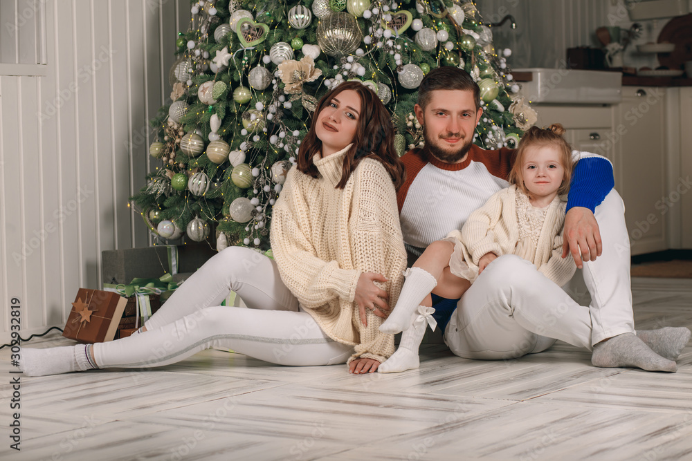Obraz premium Beautiful young family sitting next to a nicely decorated Christmas tree. Happy family having fun with Christmas presents at home. Christmas at home by the fireplace and the Christmas tree. Portrait