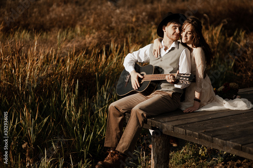 Romantic guy in a hat plays the guitar for his girlfriend on a wooden bridge near the river