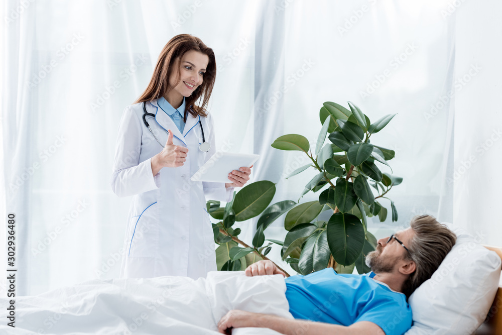 smiling doctor in white coat showing like and holding digital tablet and patient lying in bed in hospital smiling doctor in white coat showing like and holding digital tablet and patient lying in bed in hospital