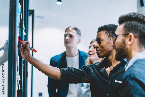Group of diverse employees concentrated on idea during meeting in office, African American men worker in formal wear writing information on sticky note while brainstorming together with colleagues
