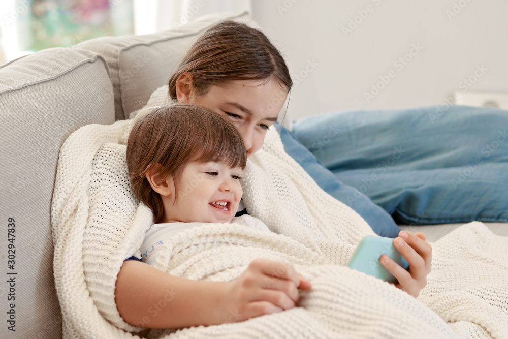 Happy siblings snuggling in warm knitted blanket at home on sofa watching cartoon laughing. Sister and brother having fun together. Autumn lifestyle, leisure activity Happy siblings snuggling in warm knitted blanket at home on sofa watching cartoon laughing. Sister and brother having fun together. Autumn lifestyle, leisure activity