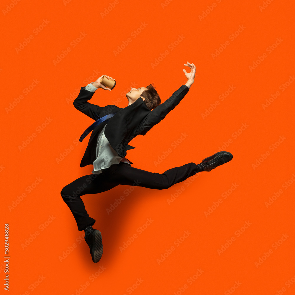 Man in casual office style clothes jumping and dancing isolated on bright orange background. Business, start-up, open-space, inspiration concept. Flexible ballet dance. Hurrying up, drinking coffee.