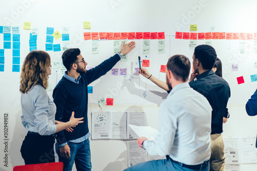 Intelligent male professional pointing on colorful stickers with text message glueded on wall and discussing information with creative multicultural colleagues having brainstorming meeting in office photo