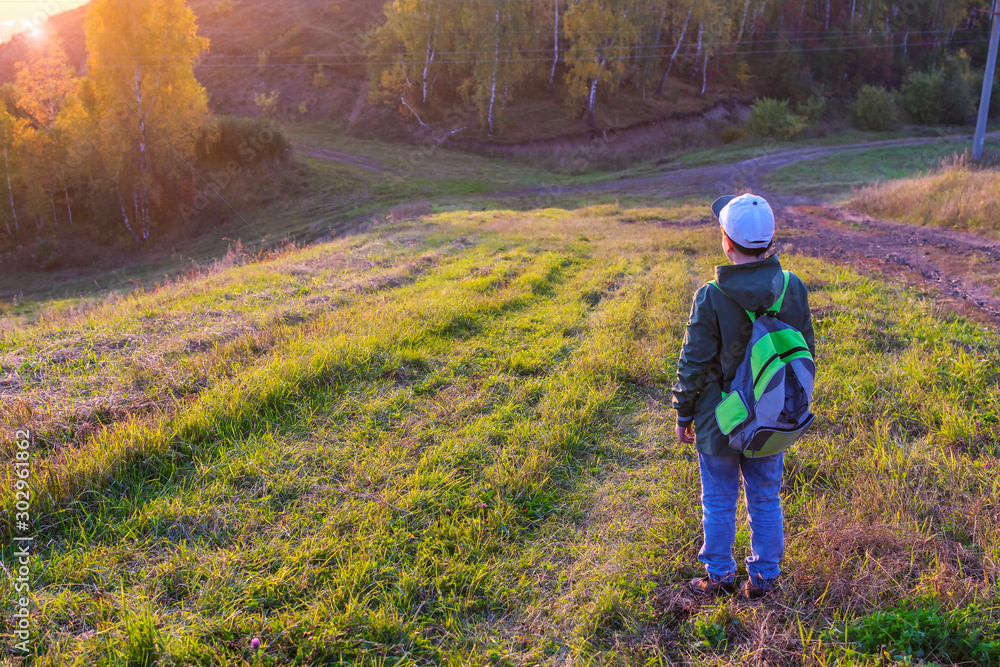 A teenage boy with a backpack stands on the trail and looks at the bright dawn. Ahead the dense forest and a fork in the road. A teenage boy with a backpack stands on the trail and looks at the bright dawn. Ahead the dense forest and a fork in the road.