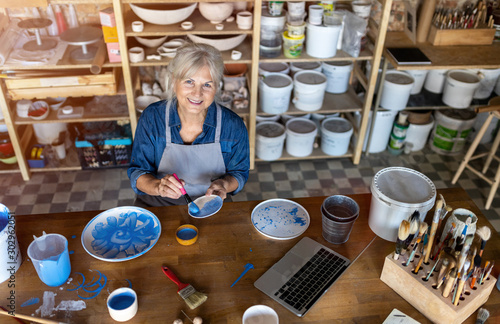 Mature craftswoman painting a plate made of clay in art studio