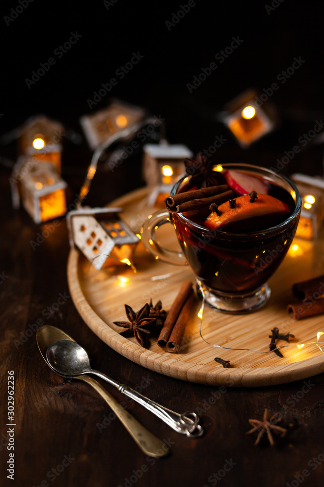 Christmas delicious hot mulled wine with orange, apple, cinnamon, clove, cardamom and anise on wooden plate. Warm beverage for cold winter days. Brights lights create festive mood. Dark background