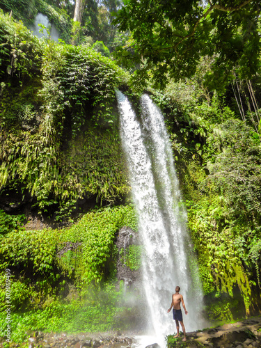 Fototapeta Naklejka Na Ścianę i Meble -  A man in swimsuit standing under two levelled waterfall in Lombok, Indonesia. Tiu Kelep Waterfall is surrounded by lush green plants from each side. Long and powerful waterfall. Beauty of the nature.