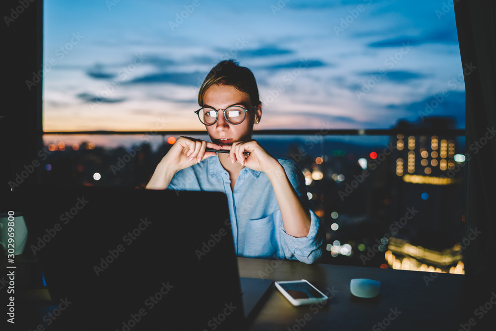 Young businesswoman dressed in shirt reading useful article about creative ideas for startup project on laptop computer.Pensive female person working overtime late at night office using modern gadget