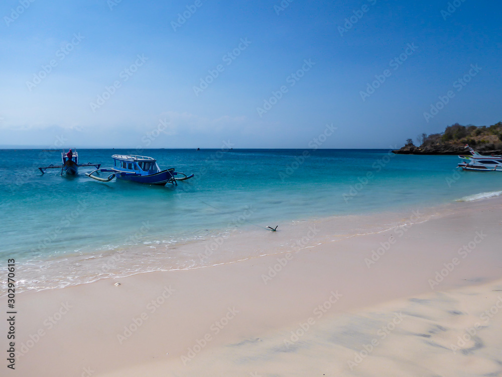A view on a bay on Pink Beach, Lombok Indonesia. A few colourful boats anchored to the shore. The water has many shades of blue. The heavenly beach is surrounded by small hills. Paradise beach.