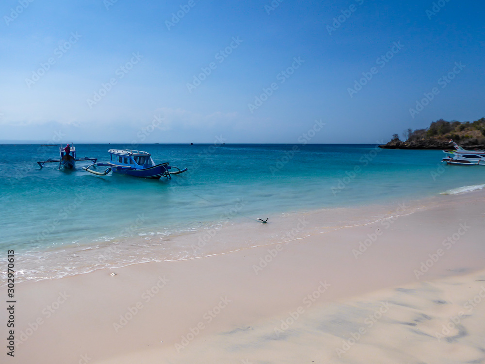 A view on a bay on Pink Beach, Lombok Indonesia. A few colourful boats anchored to the shore. The water has many shades of blue. The heavenly beach is surrounded by small hills. Paradise beach. A view on a bay on Pink Beach, Lombok Indonesia. A few colourful boats anchored to the shore. The water has many shades of blue. The heavenly beach is surrounded by small hills. Paradise beach.