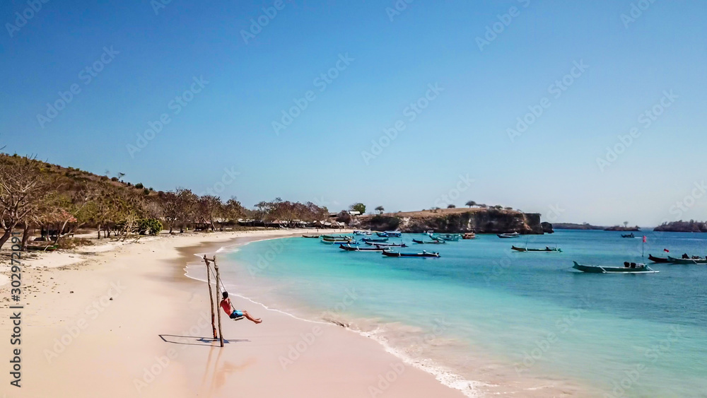 A man swinging on a swing placed on the seashore of Pink Beach, Lombok, Indonesia. The swing has simple wood construction. Waves wash the pillars of it. In the back there are few boats. Drone capture A man swinging on a swing placed on the seashore of Pink Beach, Lombok, Indonesia. The swing has simple wood construction. Waves wash the pillars of it. In the back there are few boats. Drone capture