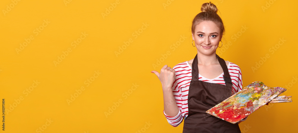 Picture of talented beautiful painter making gesture, showing direction with her thumb, holding palette in one hand, wearing dark brown apron and striped sweatshirt. Copyspace for advertisement.