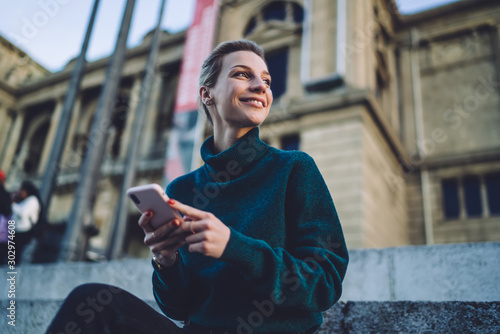 Below view of happy smiling female tourist with modern cellphone device for chatting in hands resting in city historic center and enjoying good time for getaway journey for exploring new country #302974608