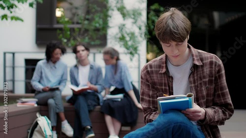 Medium shot of pensive student boy thoughtfully studying alone with group of students on background in college campus outdoor