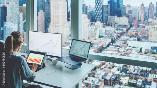 Fotografi Back view of business woman sitting at panoramic skyscraper office desktop front PC computer with financial graphs and statistics on monitor