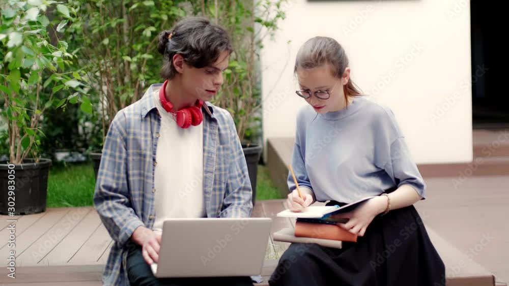 Two young casual students thoughtfully studying with book and laptop on bench in college campus outdoor Two young casual students thoughtfully studying with book and laptop on bench in college campus outdoor