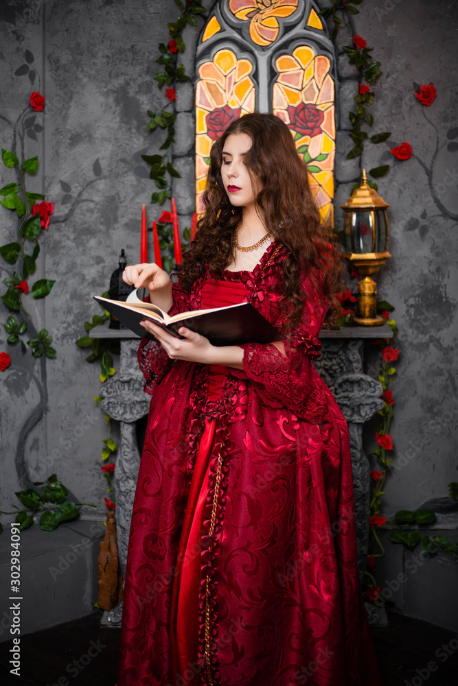 A beautiful girl in a magnificent red dress of the Rococo era stands with a book in her hands against the background of a fireplace, window and flowers.