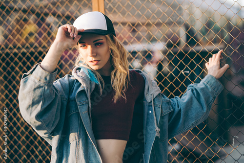 Young attractive woman in jeans jacket, shorts, red top and trucker hat posing over metal fence and graffiti wall over background in a city