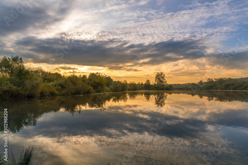 Pond at the morning sunrise over trees lying near water and moving clouds over water with reflection on water surface.