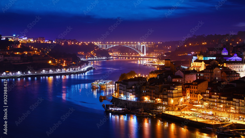 Porto, Portugal. Evening sunset panoramic view at nighttime town. Coastline of river Douro with reflections of illumination in water and picturesque clouds on blue sky.