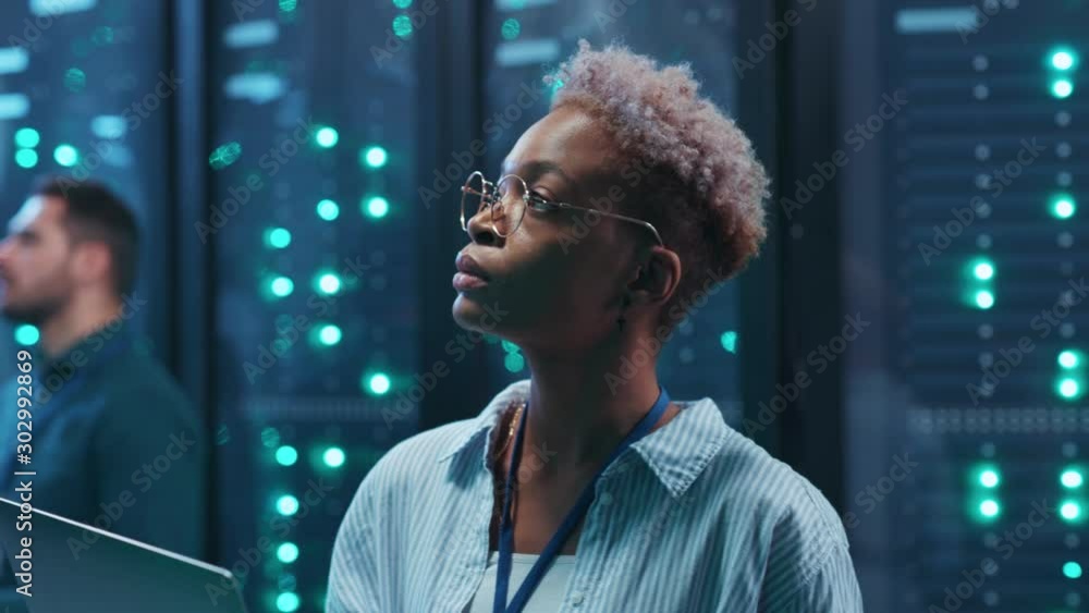 Beautiful african woman IT technician using laptop diagnosing hardware security system on network servers working in rack corridor. Close-up. Big data center.