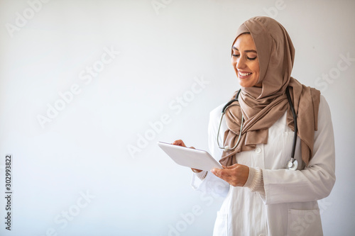 Muslimah doctor with stethoscope holding a tablet computer isolated in grey background. Confident Muslim medical student pose at hospital. Confident Muslim female doctor standing inside hospital