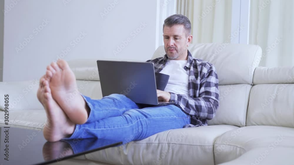 Handsome European man using a laptop computer while drinking a cup of coffee and relaxing on the sofa. Shot in 4k resolution Handsome European man using a laptop computer while drinking a cup of coffee and relaxing on the sofa. Shot in 4k resolution