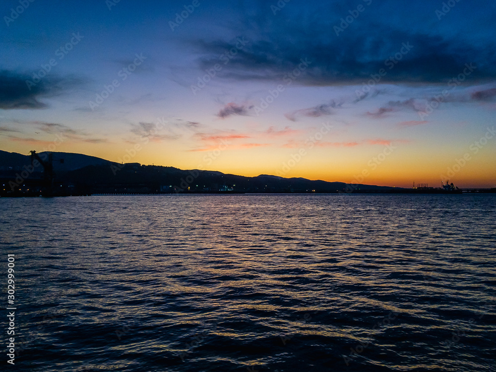 Beautiful colorful sunrise over the surface of the water of the sea with small waves with a mountainous contour of the shore of a coastal town against a blue cloudy sky.