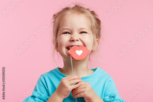 Funny smiley girl face on the background of a bright pink wall. Child girl with paper accessories, paper lips on a stick.