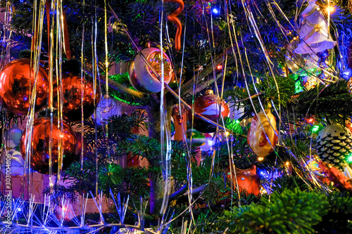 Silver tinsel on Christmas tree. Blurred holiday background with garland, glass toys, balls, lights. Colorful decoration close-up.