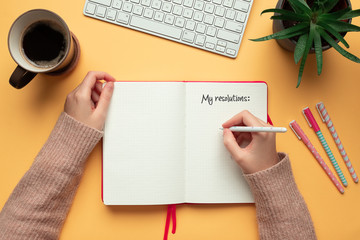 Stock photo of a young woman hands writing in a 2020 new year notebook with list of resolutions and objects on yellow background