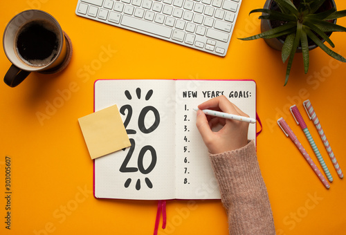 Stock photo of a young woman hand writing in a 2020 new year notebook with list of resolutions and objects on yellow background